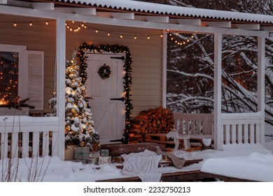 The porch and facade of the wooden white house are decorated for Christmas. The door of the cottage with a wreath is framed by a green garland. Snow-covered Christmas tree with toys and gifts. - Powered by Shutterstock