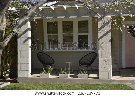 PORCH STYLED WITH CHAIRS, PASTEL PINK FRONT DOOR and beautiful leadlight windows. House is an old wooden siding period style home with a solid block supported awning, seating, lawn and potted plants