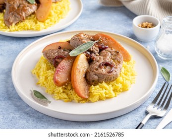 Pork chops stewed with apple slices, mustard and sage leaves served with curry rice. Food on white plate, fork and knife. Hot delicious dinner or lunch. Blue background. Close up view.