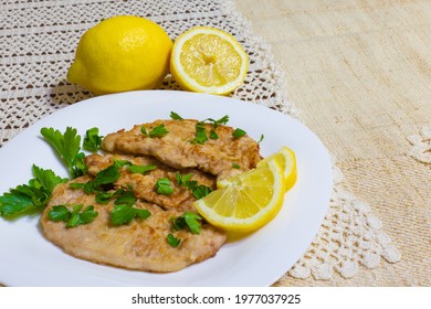 Pork escalopes in a white plate with half a lemon and parsley on an embroidered crochet tablecloth.