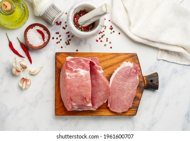 Pork. Fresh raw meat on a chopping board, salt, pepper in a mortar, garlic, olive oil and red chili pepper on a marble background. Pork chops. Selective focus, top view, copy space