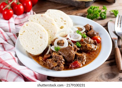 Pork goulash meat with dumplings on white plate, cutlery, garlic, onion, pepper, tablecloth in the background - typical Czech food - Shutterstock ID 2179100231
