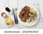 pork liver patties with fresh mixed salad and lemon slices on a white plate on white wooden table with cutlery, horizontal view from above, flat lay