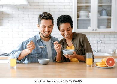 Porridge for breakfast. Weekend morning. Happy young family couple, caucasian man and african american woman, having breakfast at cozy home kitchen with healthy porridge, juice and fruits, smiling - Powered by Shutterstock