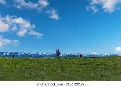 Port Angeles, WA, USA-July 2022; People Walking Near Hurricane Ridge Visitor Center With Panoramic View On The Snow Topped Mountainous Area In Olympic National Park Against A White Clouded Blue Sky 