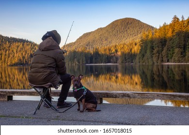 Port Moody, Vancouver, British Columbia, Canada - Feb 20, 2020: Man With A Dog Is Fishing In The Lake At White Pine Beach During A Vibrant Winter Sunset.