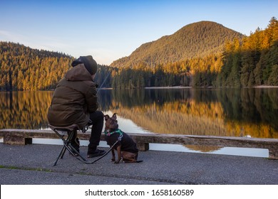 Port Moody, Vancouver, British Columbia, Canada - Feb 20, 2020: Man With A Dog Is Fishing In The Lake At White Pine Beach During A Vibrant Winter Sunset.