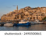 Port and old city of Jaffa illuminated by the rays of the sunset, as seen from the dock. Fishing boats in the foreground. Bell tower of the St. Peter