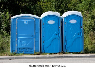 Portable blue and white ecological toilets in a row for men, woman and disabled persons next to paved road with trees and other forest vegetation in background