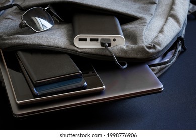 Portable charger - power bank lies in backpack along with sunglasses and gadgets - smartphones, tablet and slim fashionable laptop. Charge your gadgets while on vacation, travel and trip. Close-up