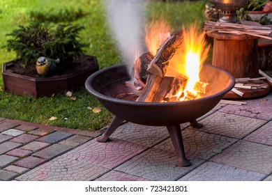 A portable fireplace with bright burning firewood making sparks and smoke at the backyard  or garden near house. A place for evening meeting and stories.