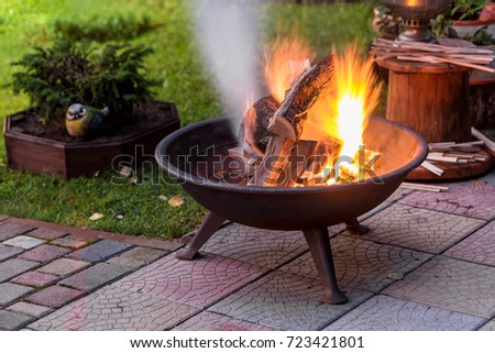 A portable fireplace with bright burning firewood making sparks and smoke at the backyard  or garden near house. A place for evening meeting and stories.