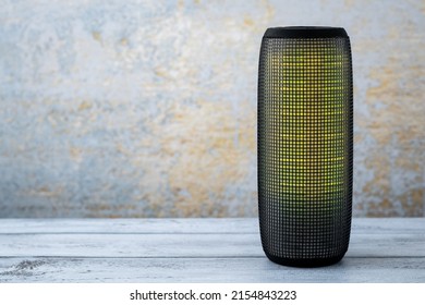 A Portable Music Column On Wooden Gray Background. Musical Wireless Speaker With Yellow Glow. Sound System. Single Object. Copy, Text Space.