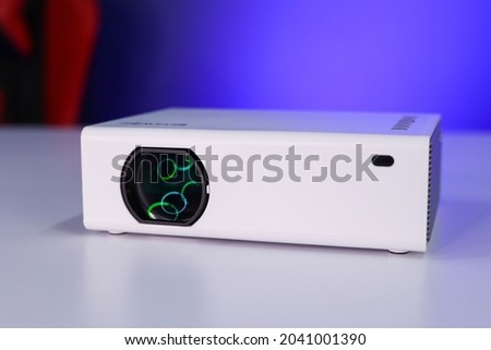 A Portable Projector on a white table in a studio, green ring light reflecting on lens. This is a Chinese projector used to watch movies and office work.