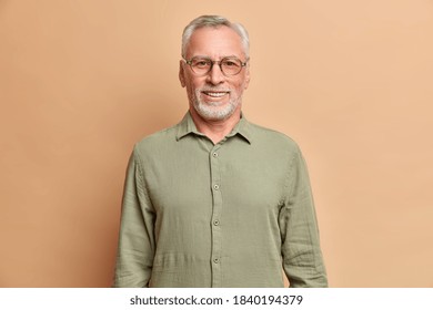 Portait of handsome European elderly man smiles positively enjoys retirement wears shirt and spectacles has perfect white teeth isolated over beige studio wall. Cheerful male pensioner indoor
