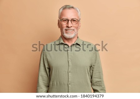 Portait of handsome European elderly man smiles positively enjoys retirement wears shirt and spectacles has perfect white teeth isolated over beige studio wall. Cheerful male pensioner indoor