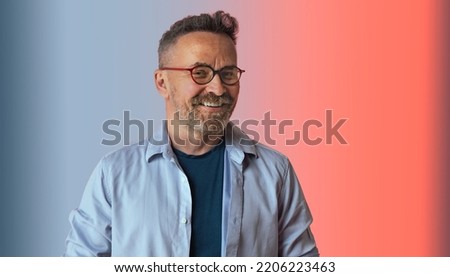 Portait of handsome European elderly man smiles positively enjoys life in blue shirt and spectacles has perfect white teeth isolated over blue pink studio wall. Cheerful mature attractive male indoor