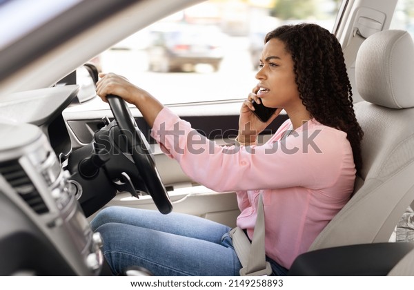 Portait of serious African American woman talking on\
mobile phone while driving car, unwary female driver using her\
cellphone, profile side view of unhappy lady arguing and riding in\
the city