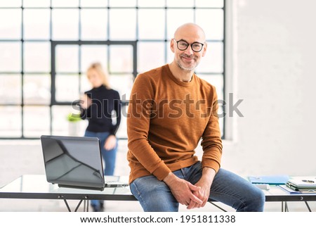 Portait shot of smiling senior businessman looking at camera and wearing casual clothes while sitting at desk in the office.
