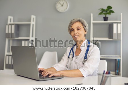 Portert positive matrue doctor woman with laptop in clinic office. A therapist with gray hair looks into the camera while sitting at a table with a computer in a hospital.