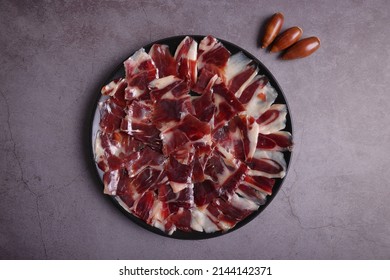 Parte de jamón ibérico de bellota 100% Dehesa de Extremadura sobre un plato negro, sobre una mesa gris oscuro y decorado con bellotas