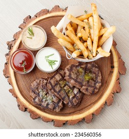 Portion Of BBQ Beef Filet Mignon Steak  Served  On Wooden Board With  Ketchup, Mustard And Cream Sauces, Fried Potatoes In Aluminium Bucket