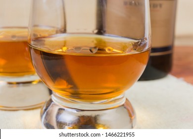 Portion of brandy in cognac bowl with drops flowing down the walls of the glass on a blurred background of other glass and bottle, fragment close-up in selective focus