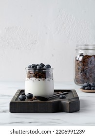 Portion Of Chocolate Granola With Yougurt Decorated Blueberry Served In A Small Jar On A Wooden Board. Big Jar With Granola On Background. Copy Space