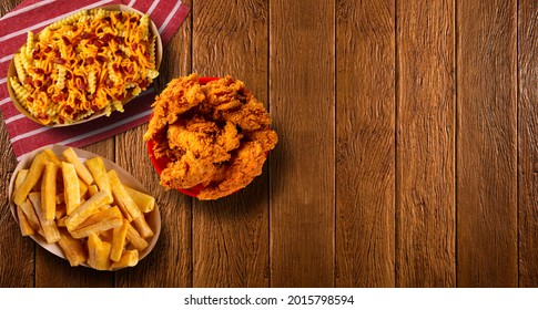 Portion Of Crispy Fried Chicken Big Red Bucket, Fried Manioc And  French Fries With Melted Cheddar Cheese And Bacon On A Wood Background.- Top View 