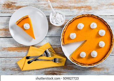 portion of delicious bright orange pumpkin open pie on plate cut from whole cake, decorated with whipped cream, with dessert forks on table napkin, close-up, top view - Powered by Shutterstock