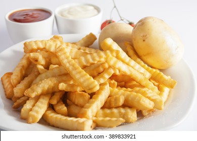 Portion of French fries (Crinkle-cut) deep fried, served on a white plate next to white bowls with mayonnaise and ketchup and fresh potato.