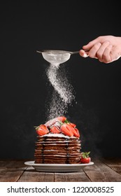 Portion Of Pancakes With Strawberries And Melted Chocolate On Black Background. Hand Sprinkling A Powdered Sugar On The Pile.