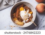 Portion of peach cobbler with a scoop of ice cream in a bowl, white marble background, top view.