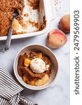 Portion of peach cobbler with a scoop of ice cream in a bowl, white marble background, top view.