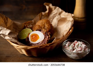 Portion Of Scotch Egg And Pico De Gallo Sauce Served On Craft Paper And Wicker Basket. Fried Pub Food Shot In The Interior Of A Bar Against A Dark Wooden Backdrop.