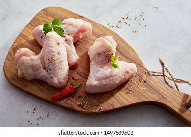 Portions Of Raw Chicken With Herbs And Spices And Red Hot Chili Peppers On A Wooden Cutting Board Over A White Marble Counter In An Overhead View