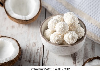 Portions of white chocolate coconut candy balls in the bowl, raw cracked coconut on the background 
