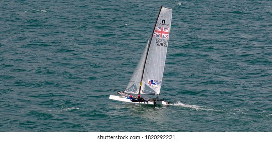 Portland Harbour, United Kingdom - July 2, 2020: High Angle Aerial Panoramic Shot Of Racing Catamaran Of The British Sailing Team. Two Sailors On It Wearing Red Helmets, British Flag On The Sails.