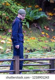 Portland, Oregon \ USA - October 28 2018: Hipster Man In Glasses With Nikon Camera On A Leather Strap Wearing Blue Jacket, Rolled Up Blue Jeans And A Grey Beanie Hat Walking In A Park