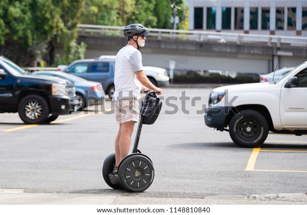 Portland, OR / USA - August 3 2018:\
Elderly person riding a black rental segway on a downtown street.\
Various cars parked in the parking lot in the\
background.