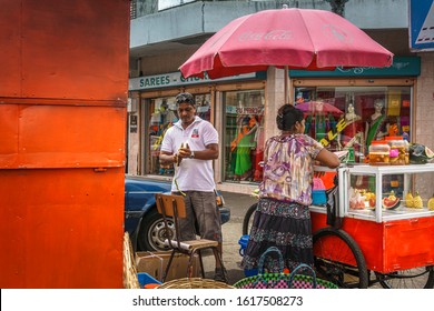 Port-Louis, Mauritius, Dec 2015 - Street Vendor Couple Selling Fruits And Pickles On The Sidewalk Facing A Saree Store