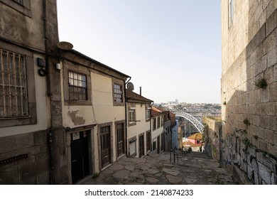Porto, Portugal. March 2022. A Typical Street Between The Old Houses Of The City Center That Goes Down Towards The River With The Dom Luís I Iron Bridge In The Background