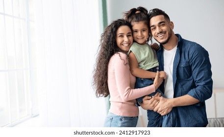 Portraif Of Happy Arabic Parents Posing With Their Little Daughter At Home, Loving Middle Eastern Family Of Three Embracing Near Window And Smiling At Camera, Enjoying Time Together, Copy Space - Powered by Shutterstock