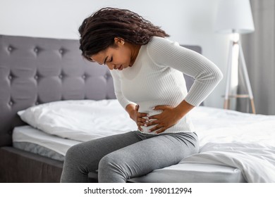 Portrair Of Upset Black Woman Suffering From Strong Abdominal Pain, Touching Her Tummy, Sitting On Bed. Sad Young Lady Feeling Acute Stomachache, Free Copy Space. Chronic Disease, Spasm, Hernia