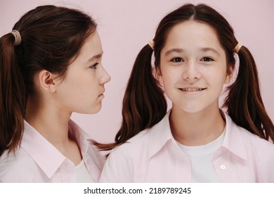 Portrait Of 12 Year Old Twin Girls. One Is Straight, Other In Profile. Both Are Wearing, Pigtails, White Shirts, Black And White Patterned Pants Over Pink Background.