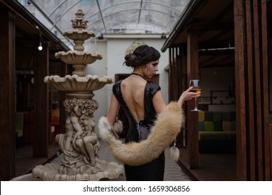 Portrait Of 20s Style Festive Beauty In A Restaurant. Young Beautiful Woman In Art-deco Style, In Black Dress In A Luxurious Interere Chicago 20s, The Era Of Gangsters, With A Glass At The Fountain