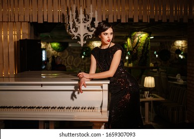 Portrait Of 20s Style Festive Beauty In A Restaurant. Young Beautiful Woman In Art-deco Style, In Black Dress In A Luxurious Interere Chicago 20s, The Era Of Gangsters. Singer  At The White Piano