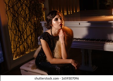 Portrait Of 20s Style Festive Beauty In A Restaurant. Young Beautiful Woman In Art-deco Style, In Black Dress In A Luxurious Interere Chicago 20s, The Era Of Gangsters. Singer  At The White Piano