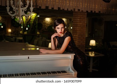 Portrait Of 20s Style Festive Beauty In A Restaurant. Young Beautiful Woman In Art-deco Style, In Black Dress In A Luxurious Interere Chicago 20s, The Era Of Gangsters. Singer  At The White Piano