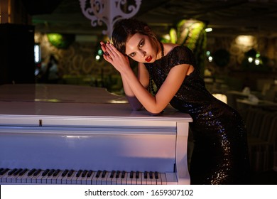 Portrait Of 20s Style Festive Beauty In A Restaurant. Young Beautiful Woman In Art-deco Style, In Black Dress In A Luxurious Interere Chicago 20s, The Era Of Gangsters. Singer  At The White Piano